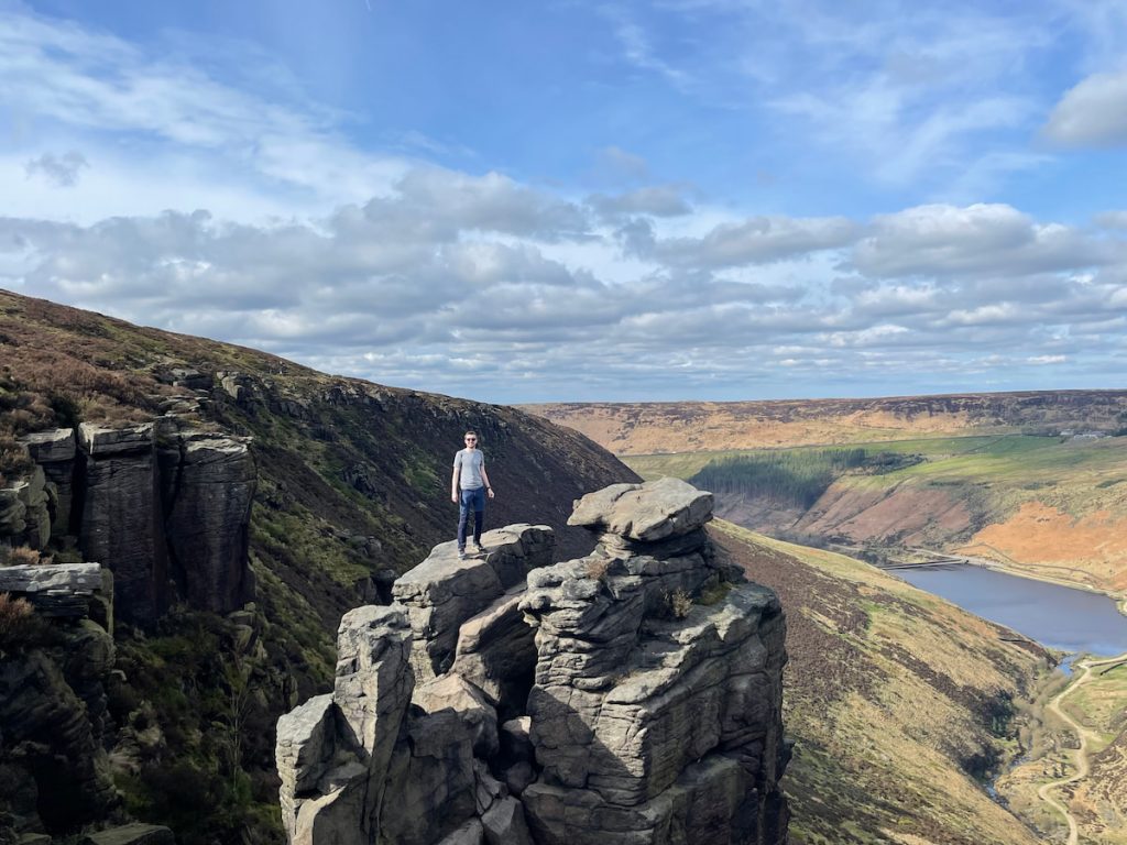 alex hesp-gollins standing on top of the Trinnacle; with a view over Greenfield reservoir below